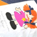 Desenho de menina feito de materiais. Cabelo de lã cor de laranja e laços azuis, um em cada lado. Vestido cor de rosa com uma estrela amarela no peito e sapatos pretos.
